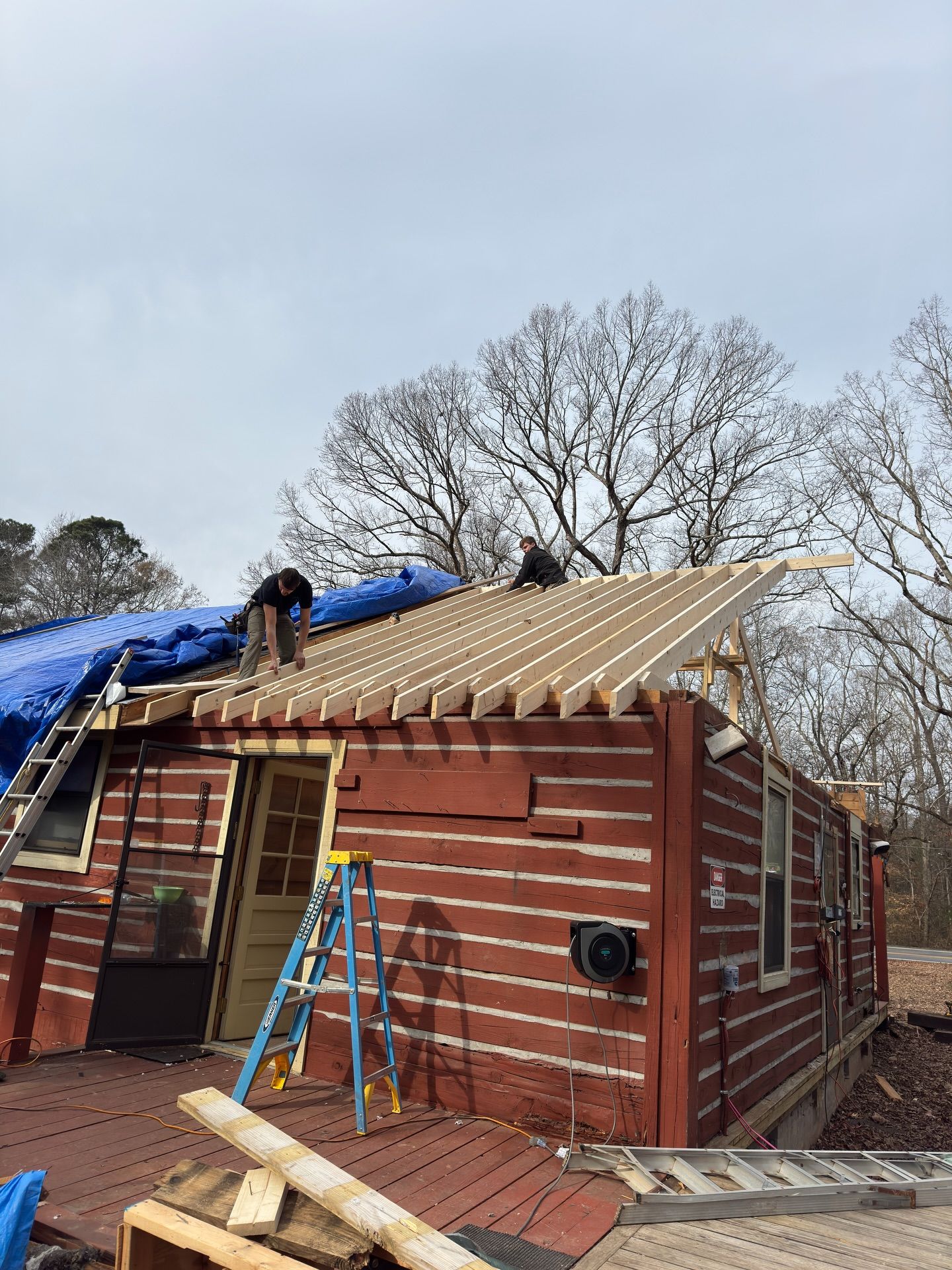 Two workers in red shirts install wooden roof rafters on a rustic, red-sided building outdoors under a cloudy sky.