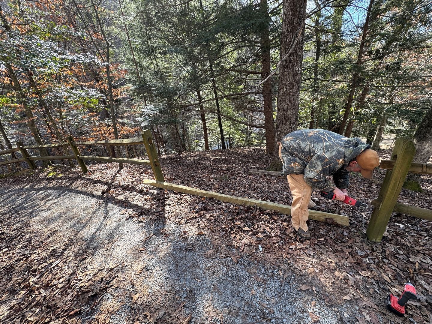 A person in camouflage gear uses a power drill to install a wooden rail on a fence along a forest trail.
