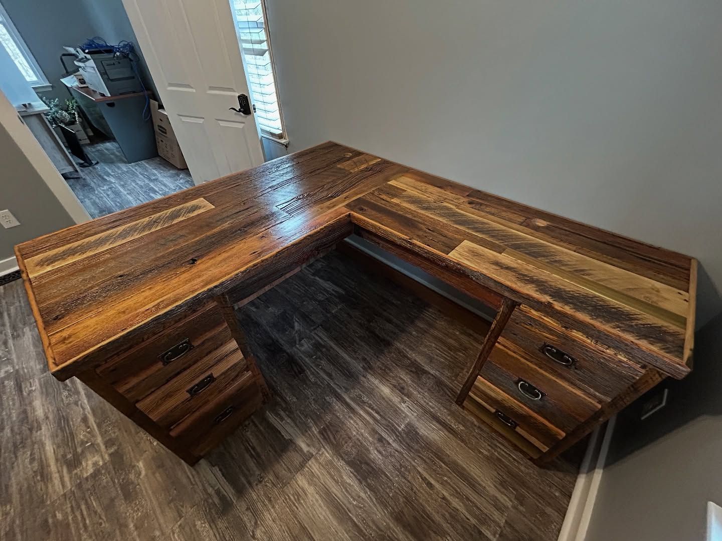 An L-shaped wooden desk with a rustic finish, featuring two built-in drawer units, set in a room with light gray walls.