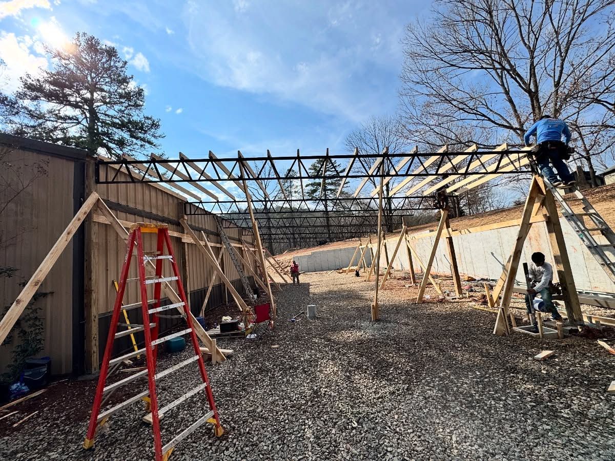 Construction workers install steel roof trusses on a wooden structure at an outdoor, sunlit job site.