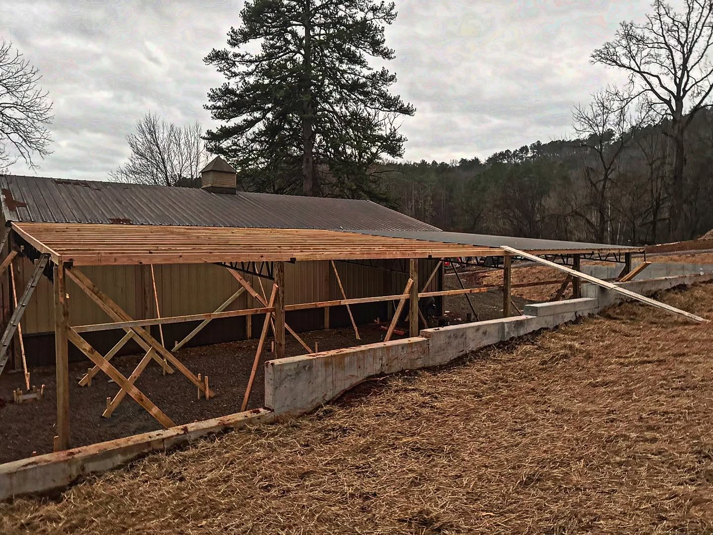 A wooden open-frame shed addition is under construction beside a barn, with a concrete foundation on sloping ground.