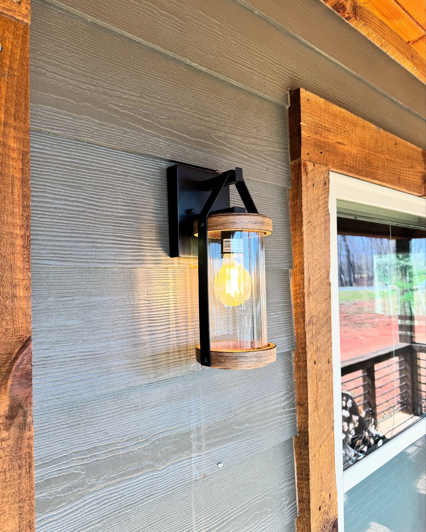 A modern farmhouse-style outdoor wall light with a glass cylinder, black metal frame, and wood accents on a gray siding.