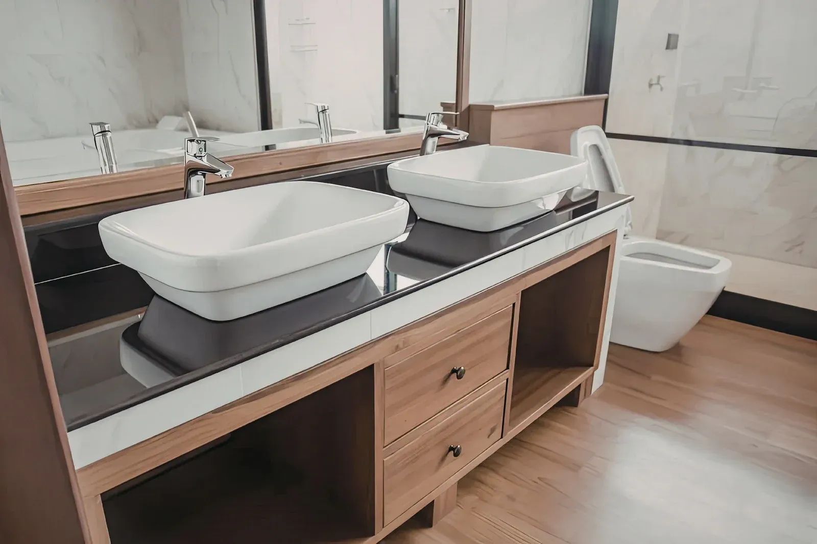Modern bathroom vanity with dual white vessel sinks, wooden cabinetry, and a white toilet against a marble-tiled wall.