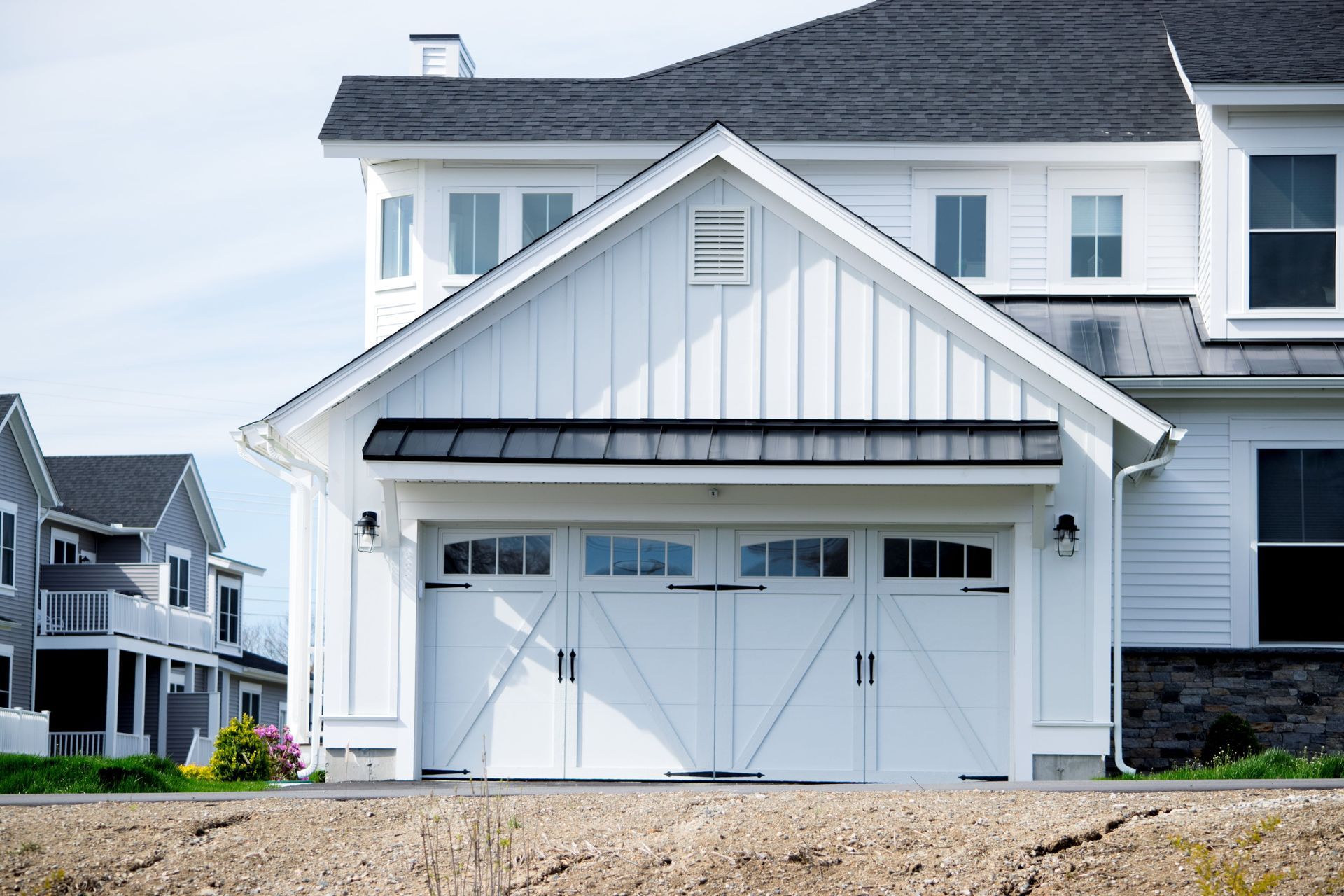 White house with a barn-style garage, black roof, and windows. Other houses visible in the background.
