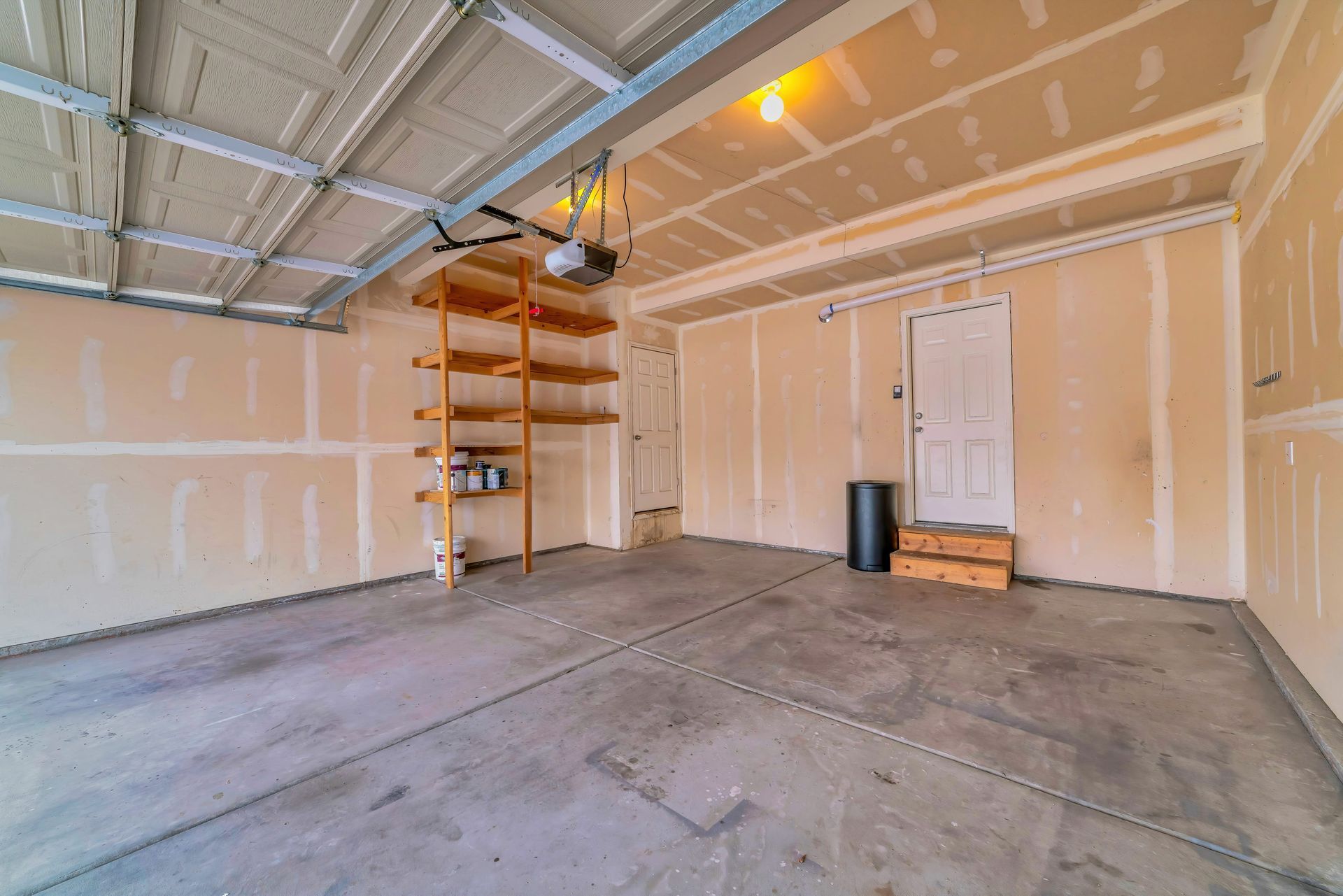 Empty garage with concrete floor, drywall walls, and a wooden shelf unit.