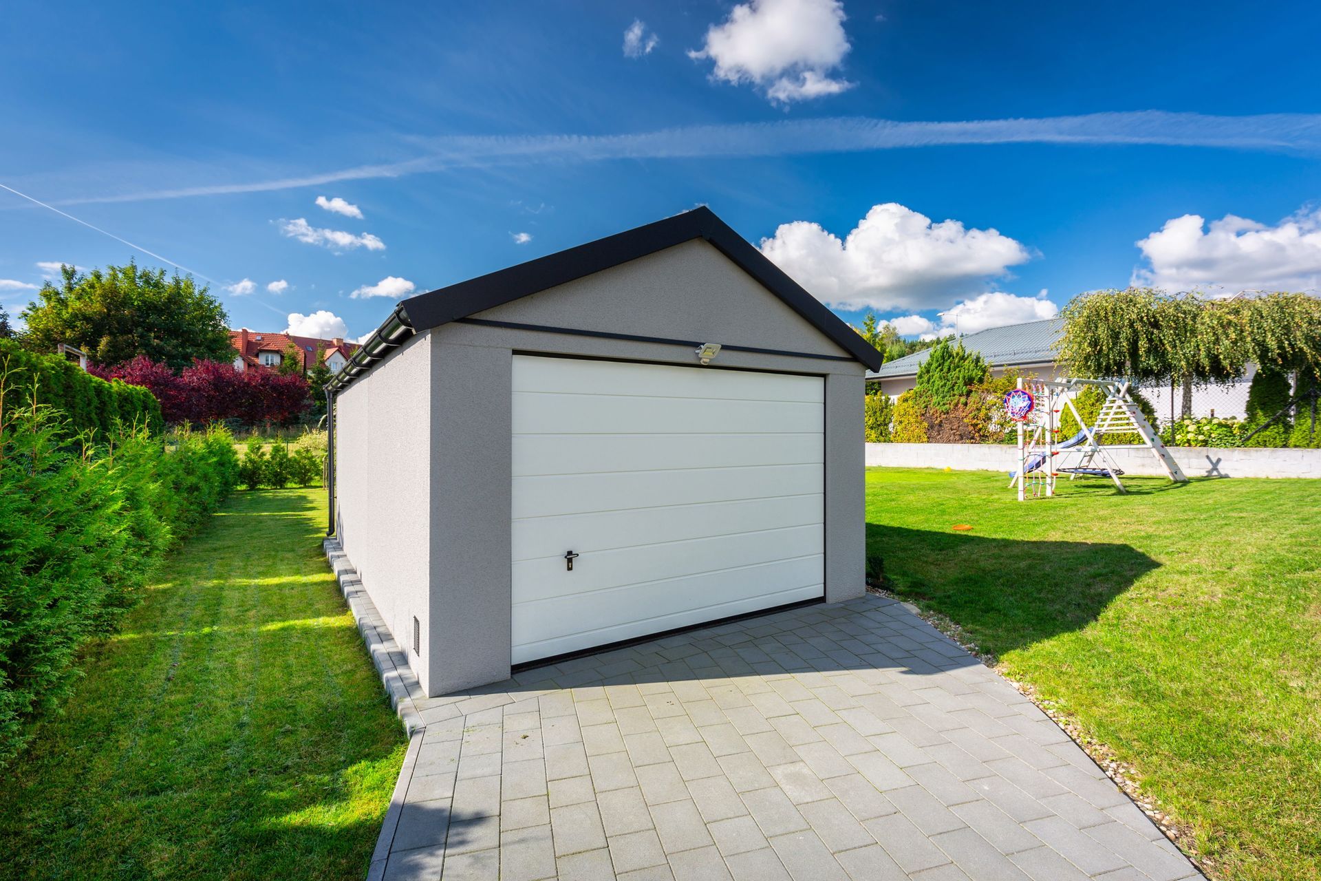Gray garage with white door and paved driveway, set on a green lawn with blue sky and clouds.