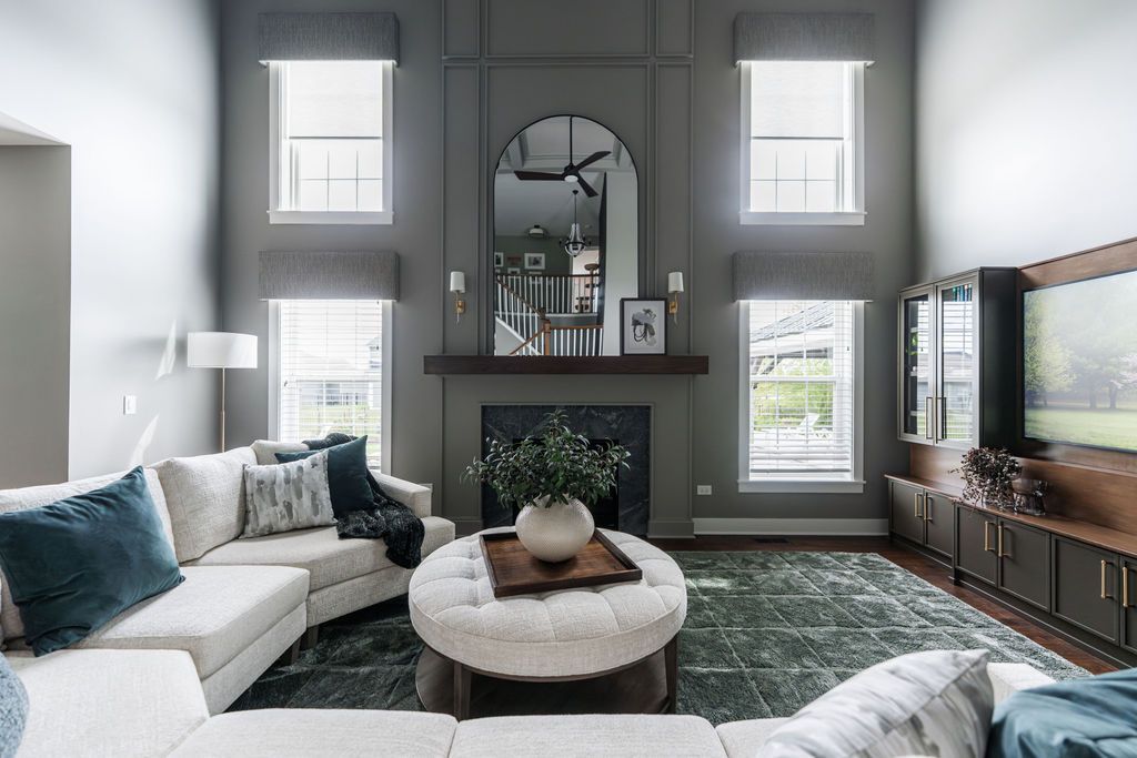 Bright modern living room with white sofas, round ottoman, large rug, and a fireplace beneath an arched mirror.