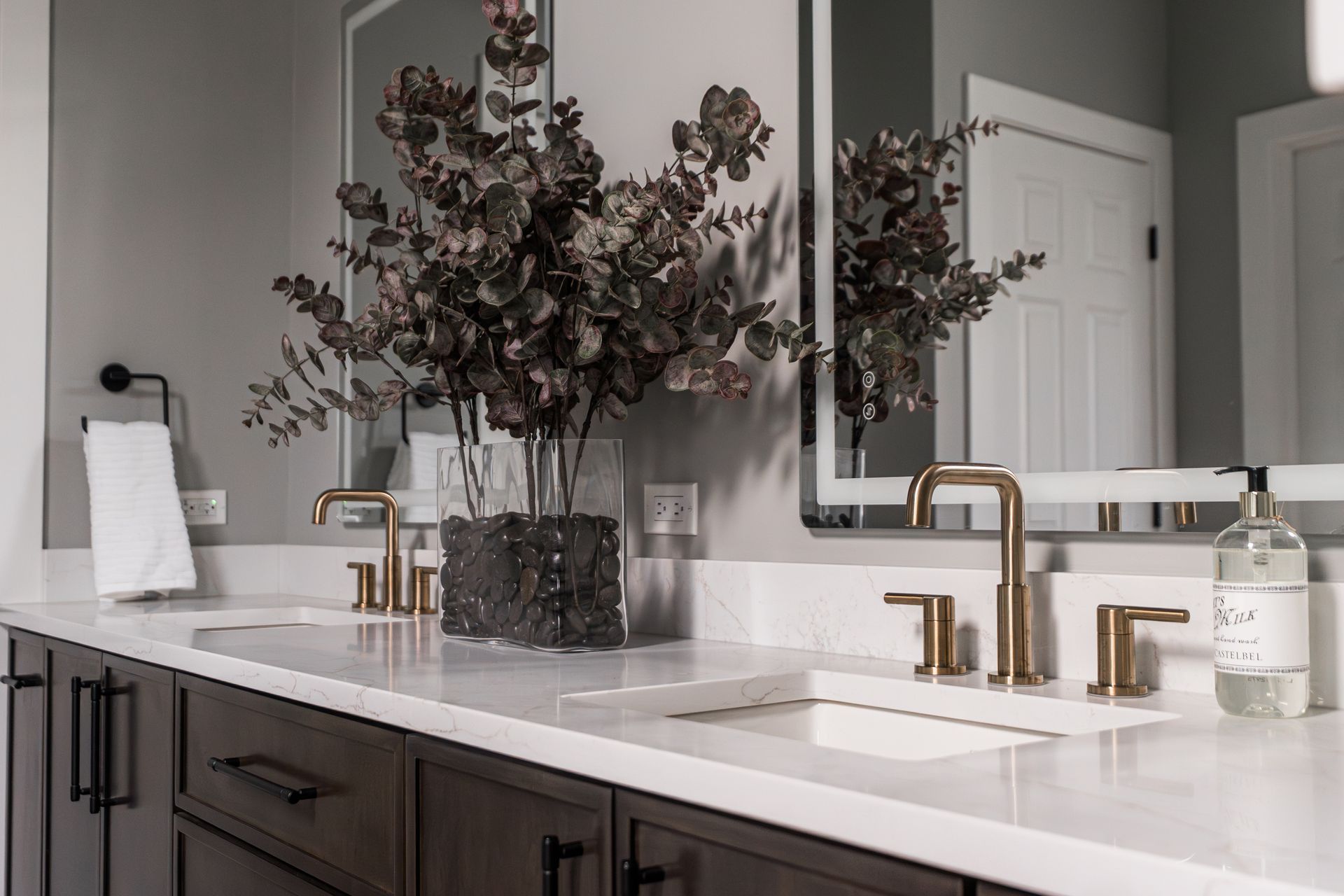 Modern bathroom vanity with double sinks, brass faucets, large mirror, and vase of dark foliage