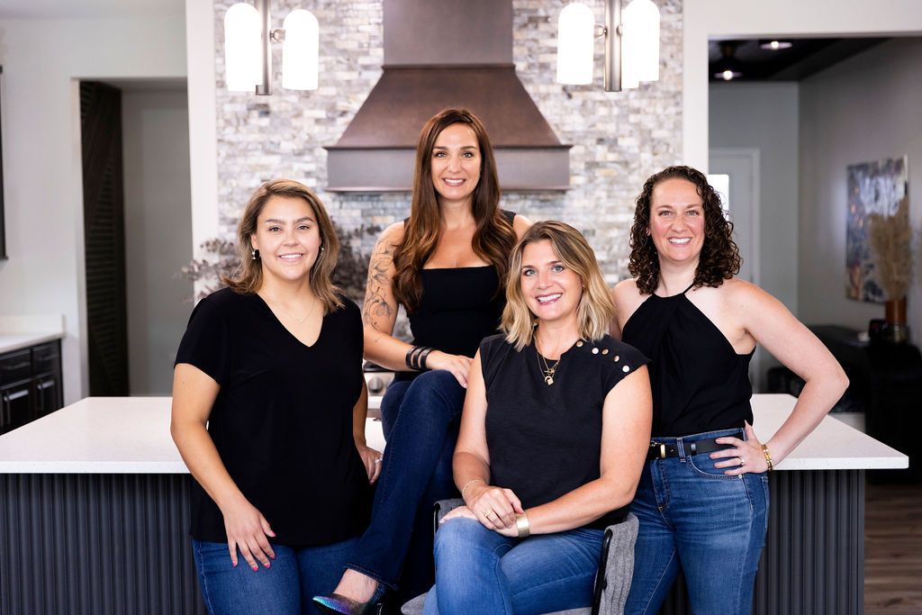 Four women posing in a modern kitchen. They wear black tops and jeans, smiling, with a white countertop and decorative backsplash.