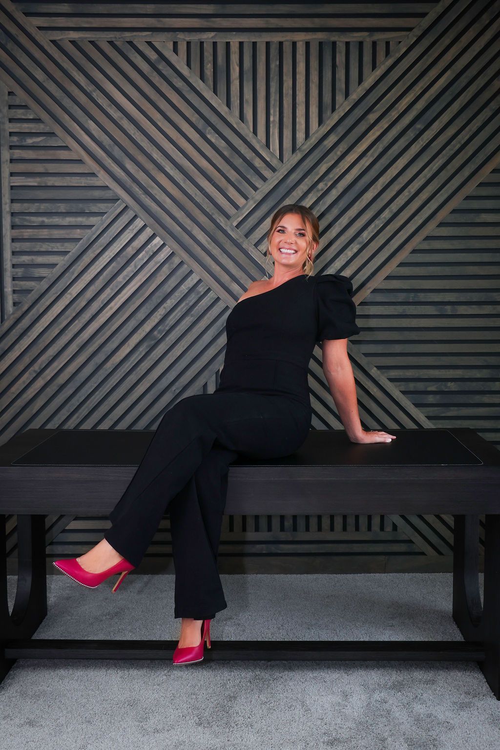 Woman in black outfit and pink heels sits on a dark bench, smiling. Behind her, a textured wooden wall.