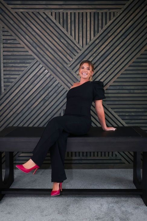 Woman in black outfit and pink heels sits on a dark bench, smiling.  Behind her, a textured wooden wall.