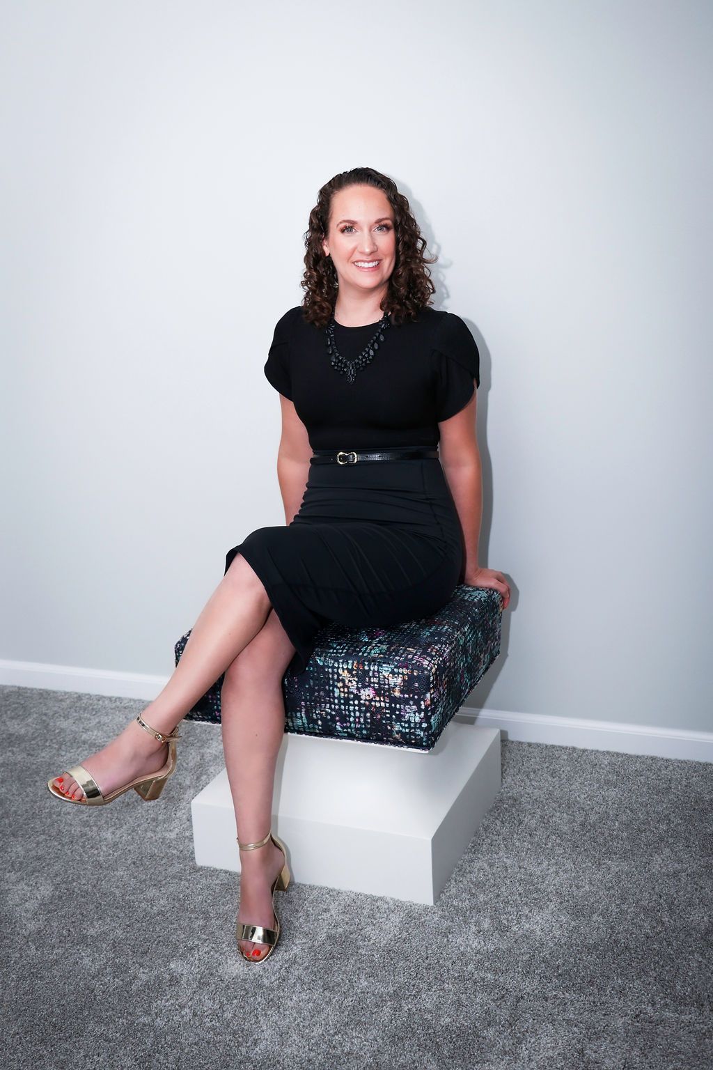 Woman in black outfit sits on a patterned cushion, smiling. Gold heels. Light background.