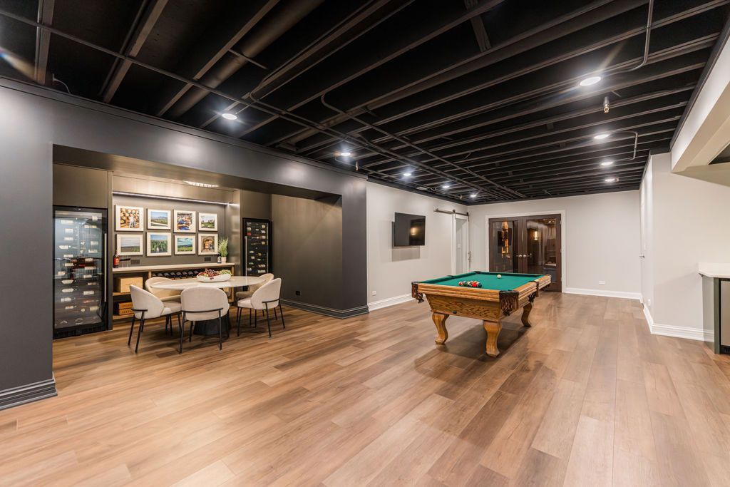 A spacious basement recreation room with a pool table, wooden floors, dining area, and exposed dark-painted ceiling.