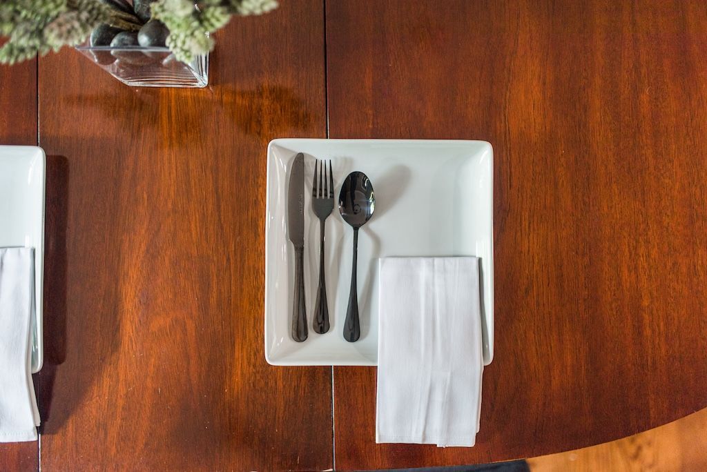 Place setting on a brown wooden table with square white plate, black silverware, and white napkin.