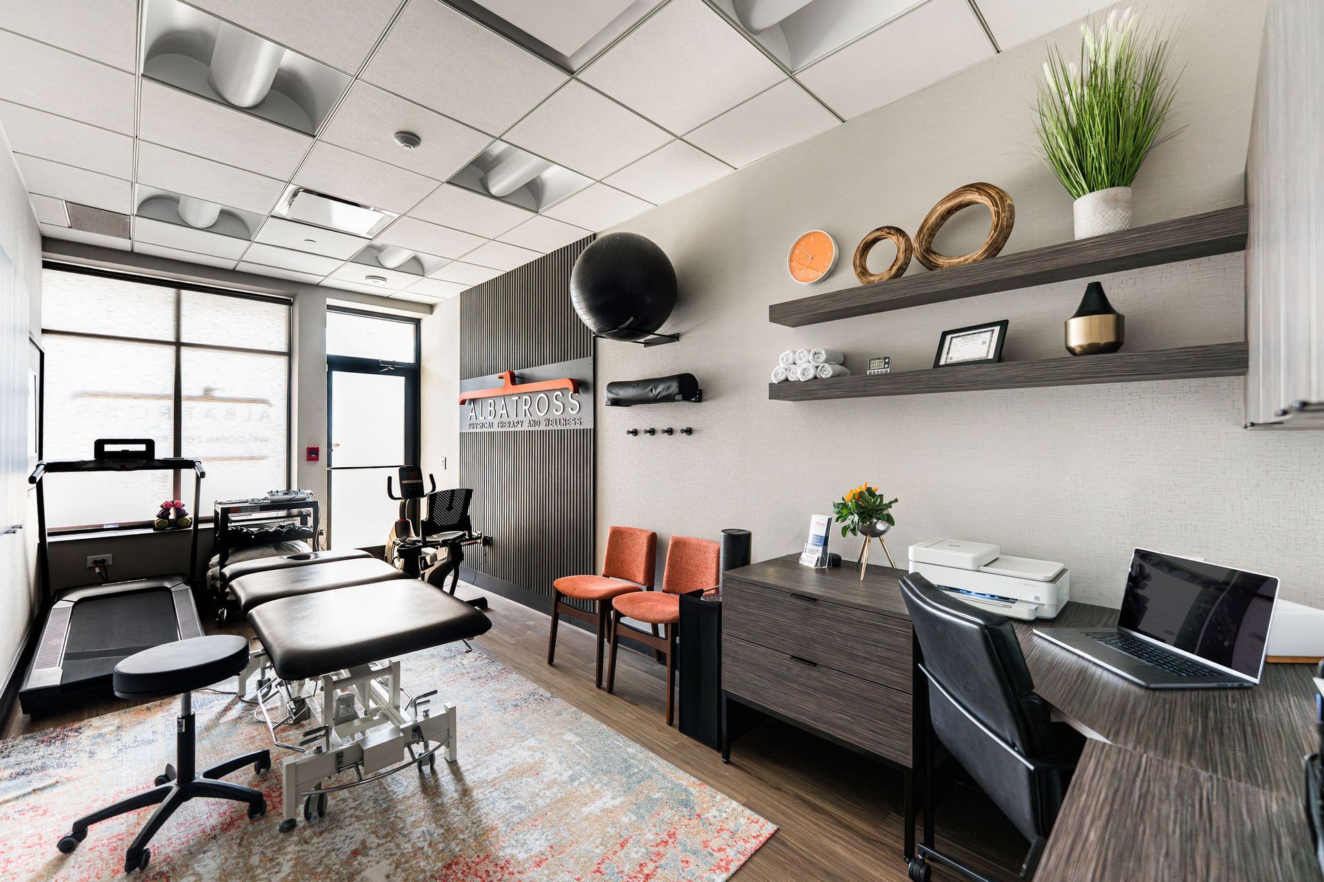 Chiropractor's office: treatment table, treadmill, chairs, shelves with decor, desk with computer and printer.