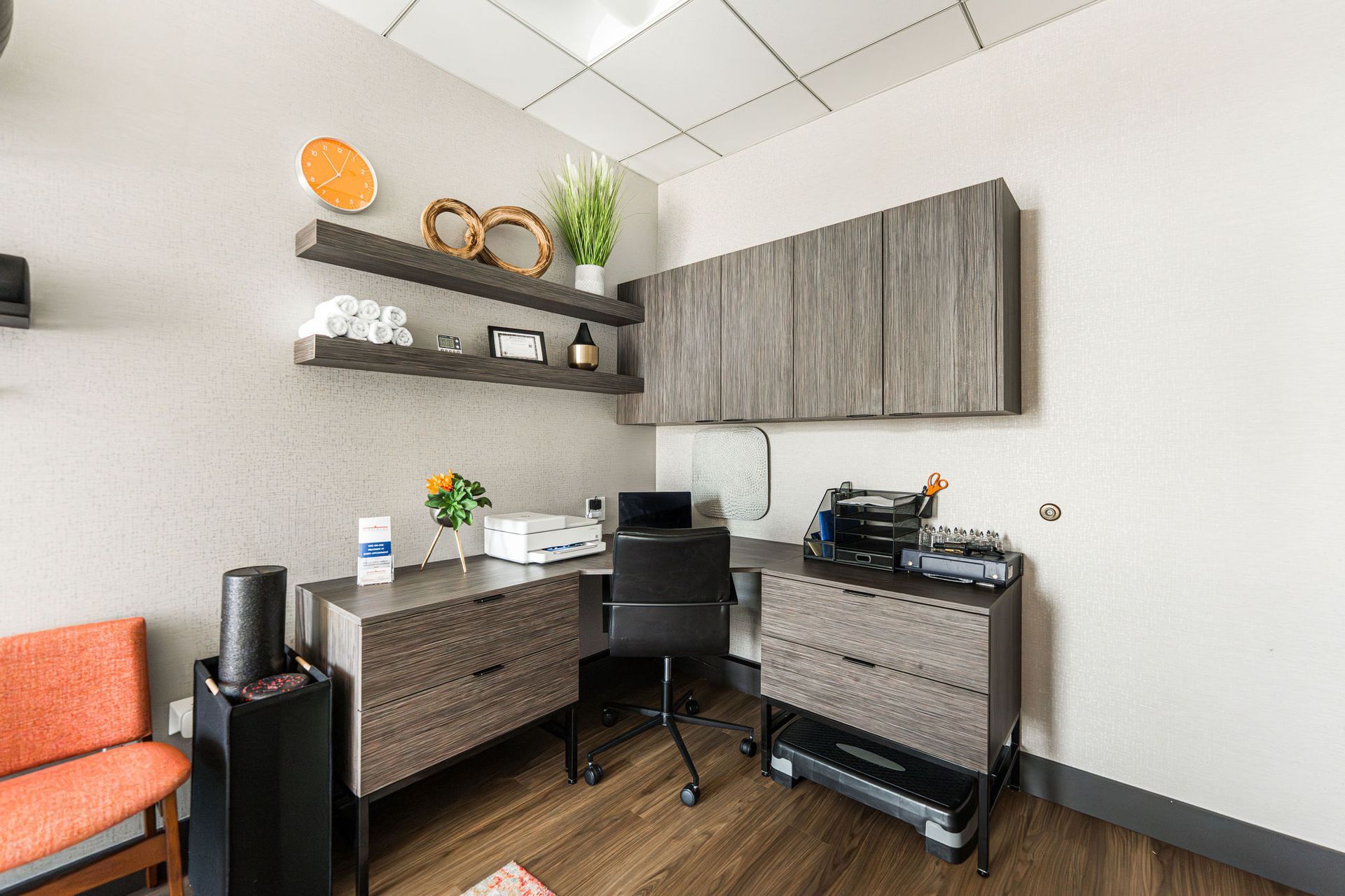 Office corner with desk, shelves, and orange chair. Light wood and gray cabinets, white walls, and wood floor.