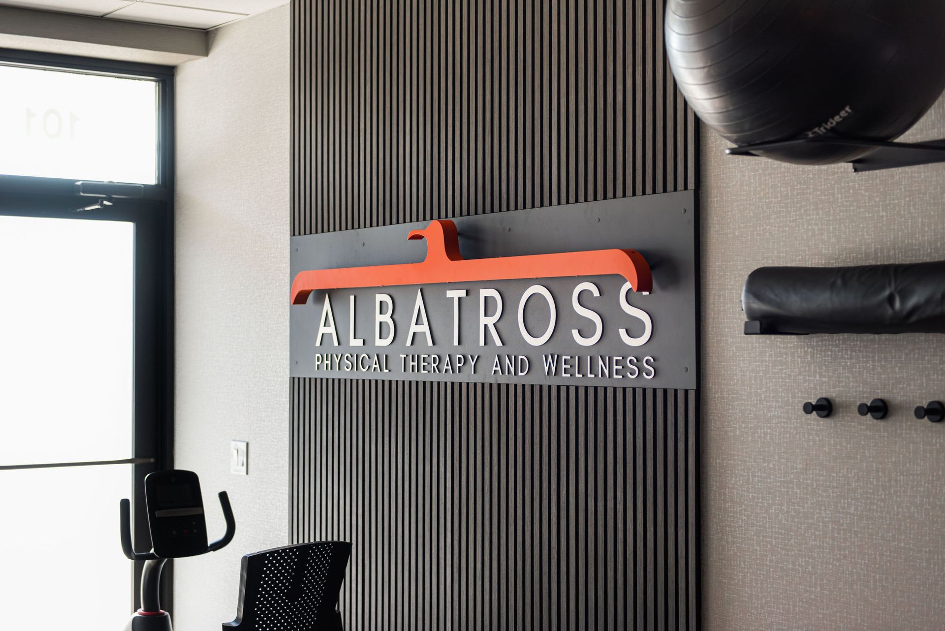 Sign for Albatross, Physical Therapy and Wellness, with orange logo on a gray wall.