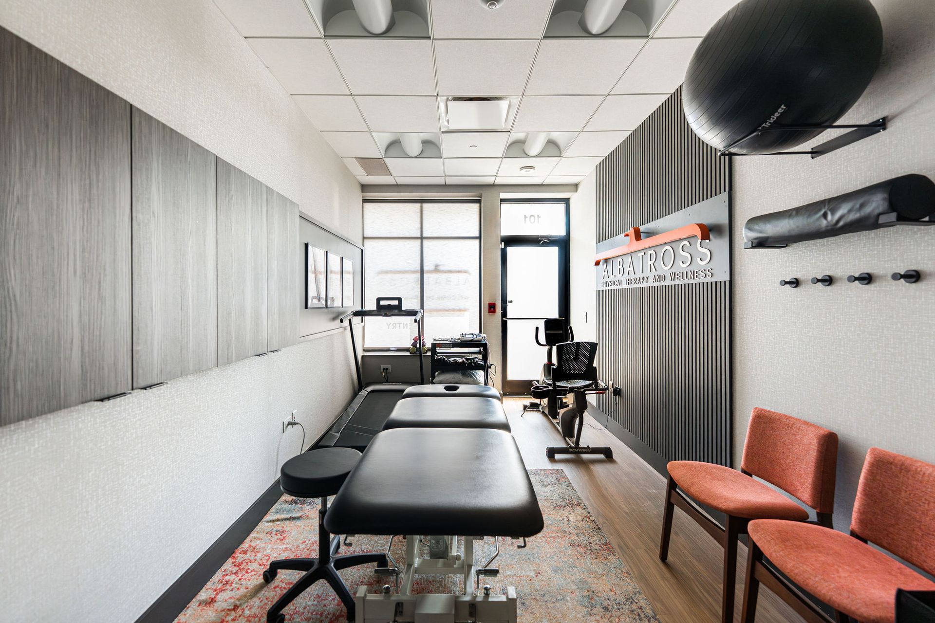 Rehabilitation room with a massage table, exercise equipment, gray walls, and orange chairs.