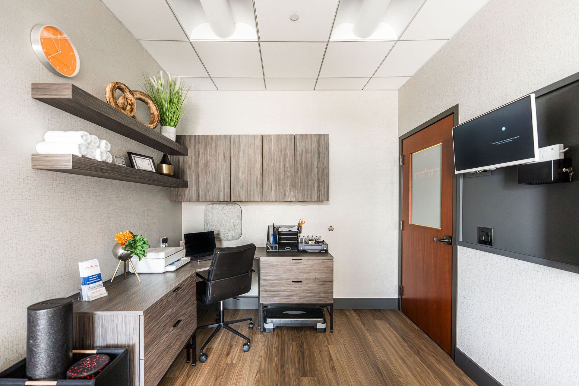 Office with a desk, shelves, and wood-look flooring. A monitor hangs on a wall.