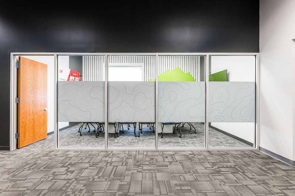 Office space with glass wall panels and frosted privacy strips; a meeting table is visible inside.