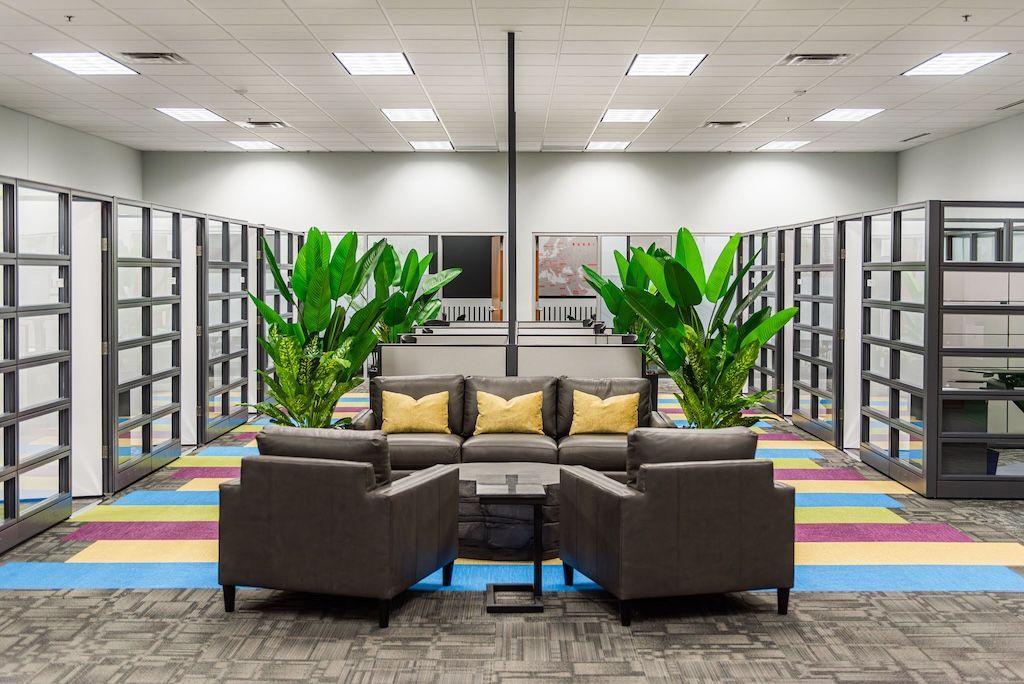 Office lounge area with sofa, armchairs, plants, and colorful carpet. Bookshelves line the walls.