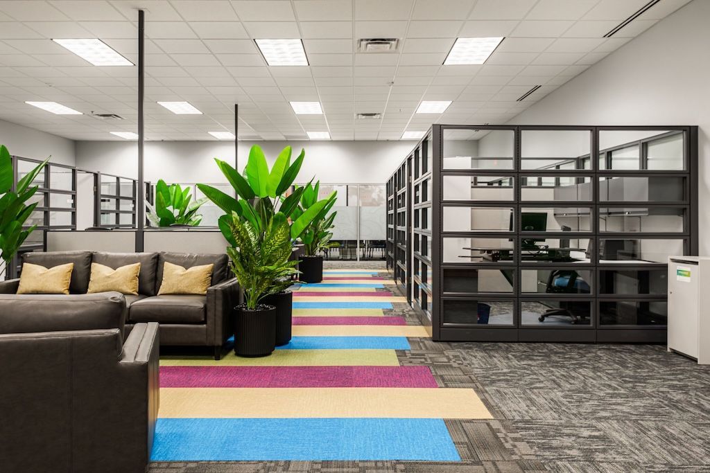 Office interior with lounge area, colorful floor, plants, and glass-walled workspaces.