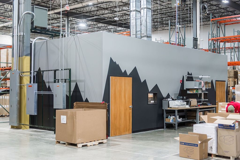 Large warehouse with a gray, mountain-decorated enclosure. Boxes are in front, with a person visible on the right.