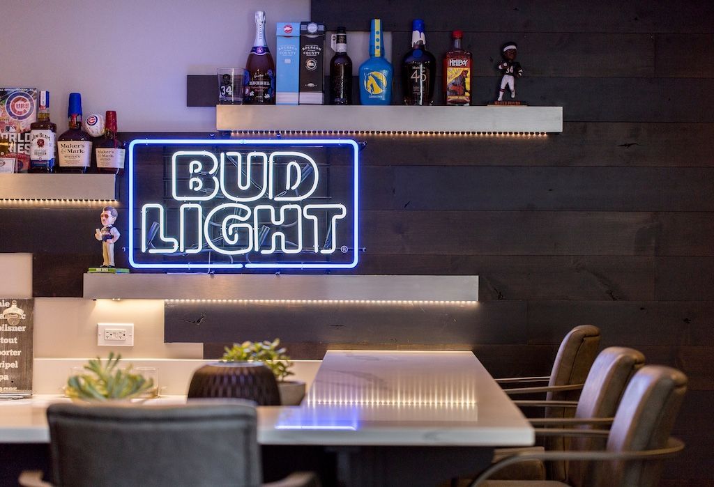 Bar with Bud Light neon sign, liquor bottles, and stools, with gray and wood paneling.