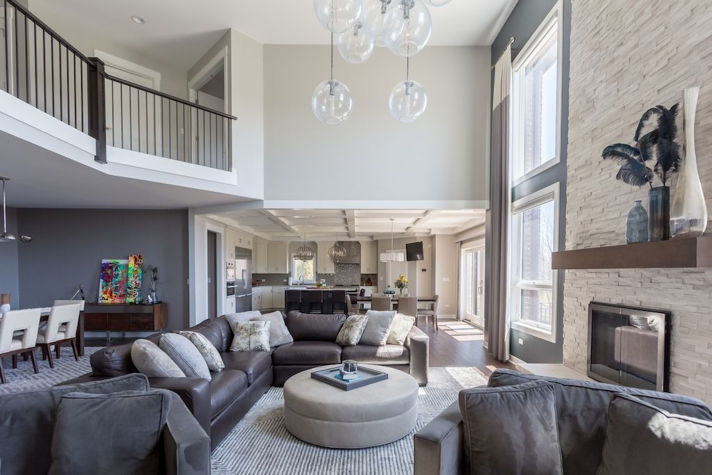 Spacious living room with high ceilings, fireplace, and open balcony. Features gray sofas, round ottoman, and glass pendant lights.