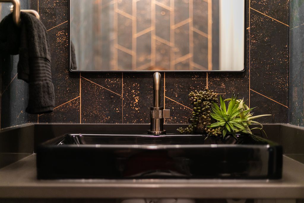Black rectangular bathroom sink with bronze faucet and a succulent. Dark patterned wallpaper and mirror.