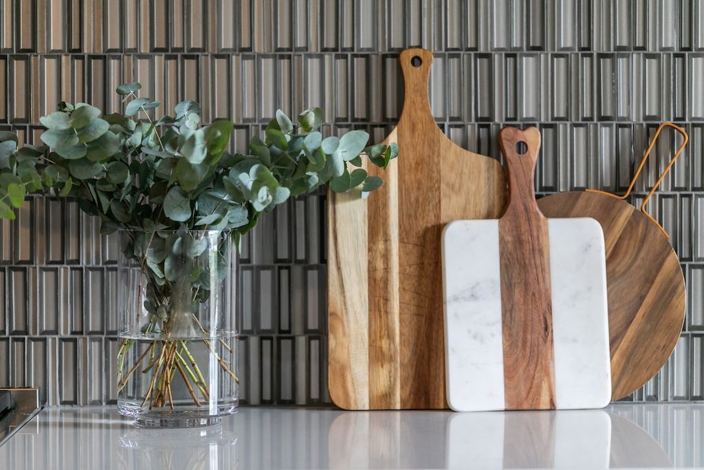 Glass vase with eucalyptus next to wooden cutting boards on a countertop, against a tiled wall.
