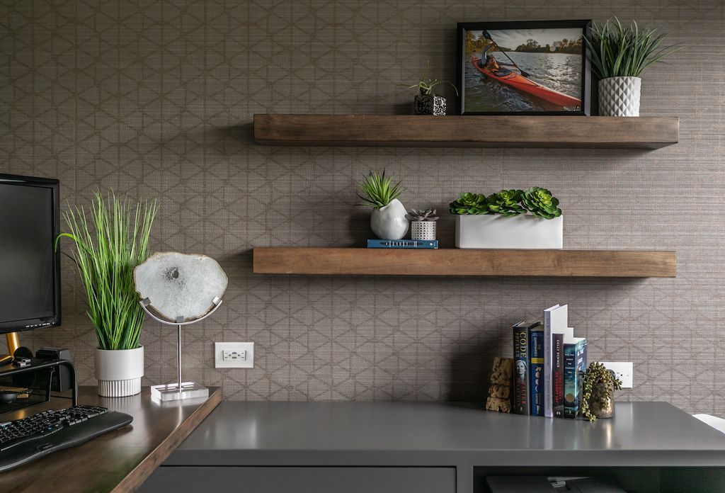 Two wooden shelves with plants and décor on a gray textured wall above a gray desk with a computer and keyboard.