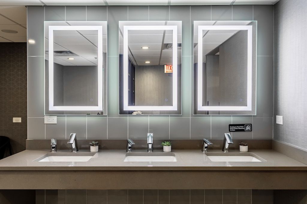 Three illuminated mirrors above sinks in a public restroom. Gray walls, countertops and tiling.