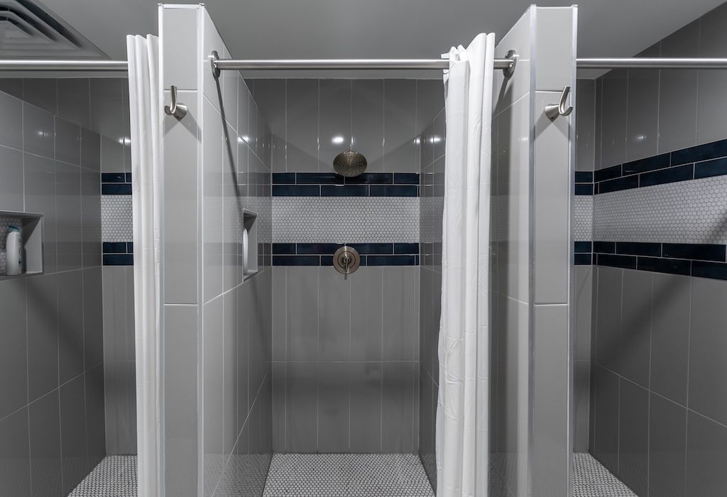 Three shower stalls with gray tile and blue accents. White shower curtains hang.