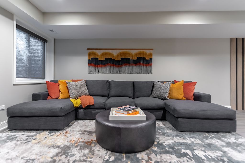 Gray sectional sofa with colorful pillows, round ottoman, and abstract rug in a modern living room.