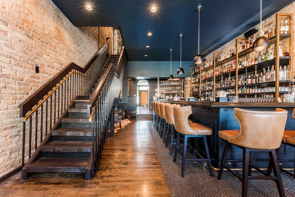 Interior of a bar with a wooden staircase and a long bar with stools. Exposed brick wall and dark blue ceiling.