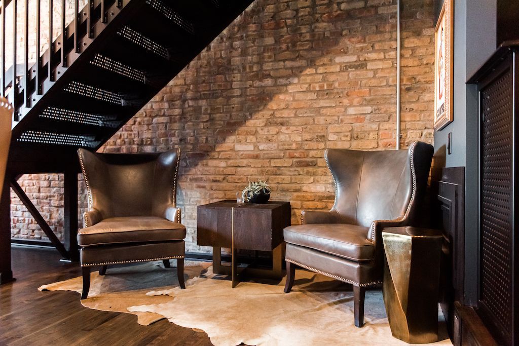 Two leather armchairs sit on a cowhide rug in front of a brick wall under a metal staircase.
