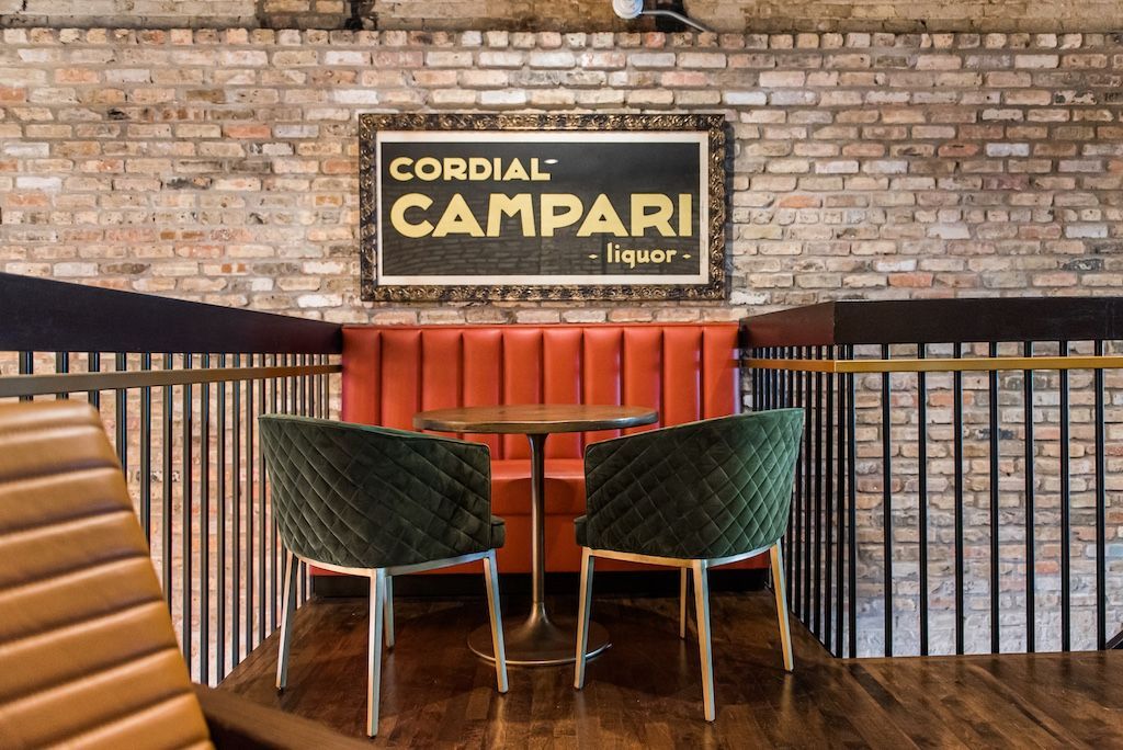 Cozy bar seating with red booth, green velvet chairs, small table, and Campari sign on brick wall.