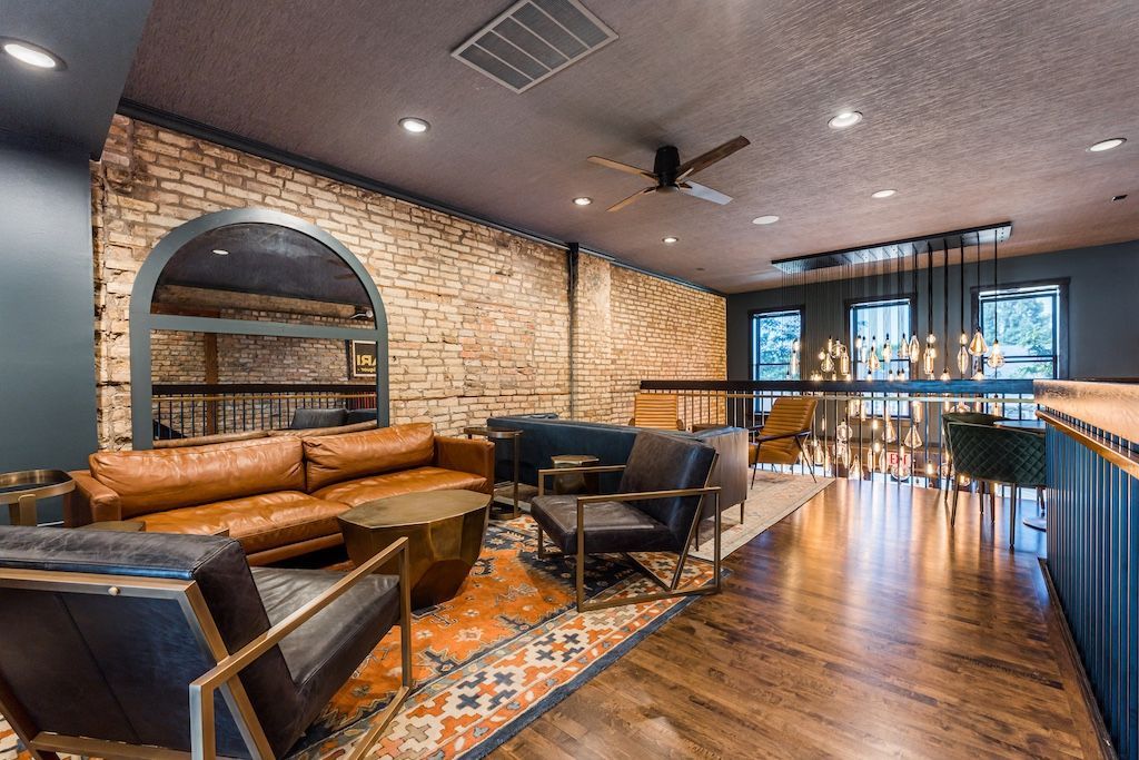 Lounge area with exposed brick, brown leather sofas, patterned rug, wooden floors, and modern lighting.