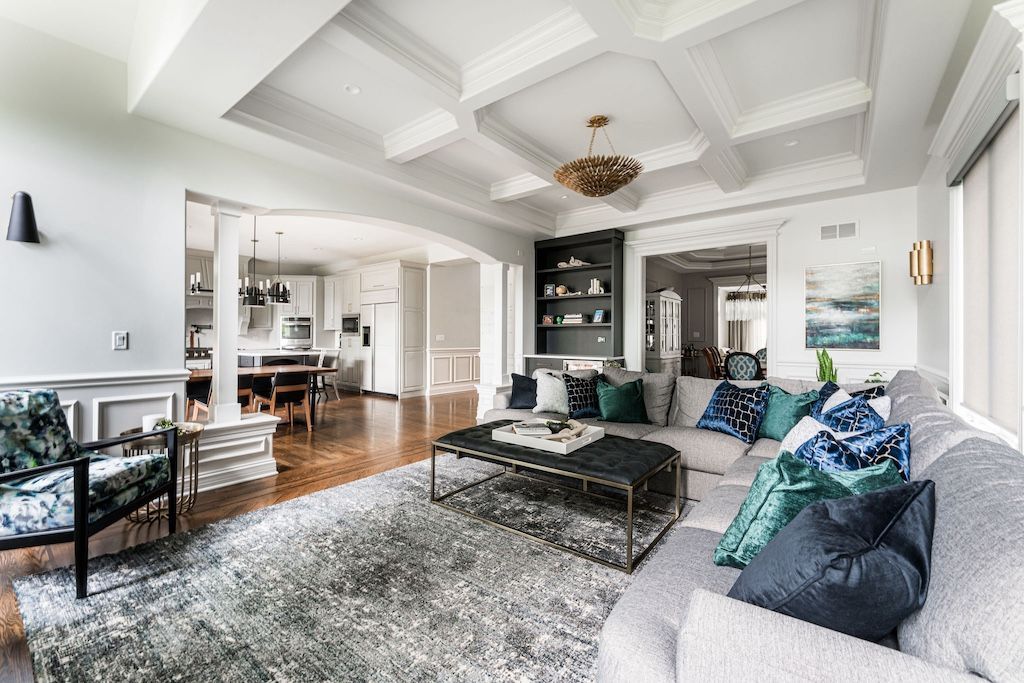 Spacious living room with white walls, coffered ceiling, open floor plan. Beige couch, patterned rug, open to dining area.