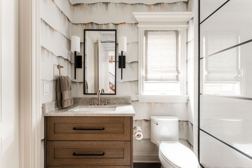 Bathroom with wood vanity, textured wall, black mirror and sconces, toilet, and window with blinds.