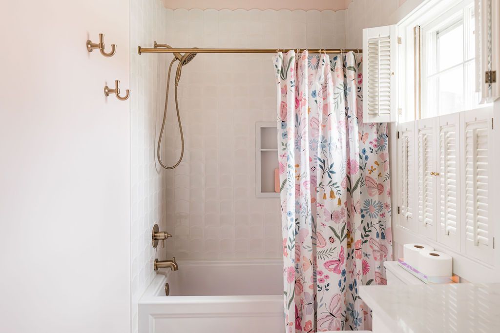 Bathroom with white walls and tub, floral shower curtain, and open shutters; brass fixtures.