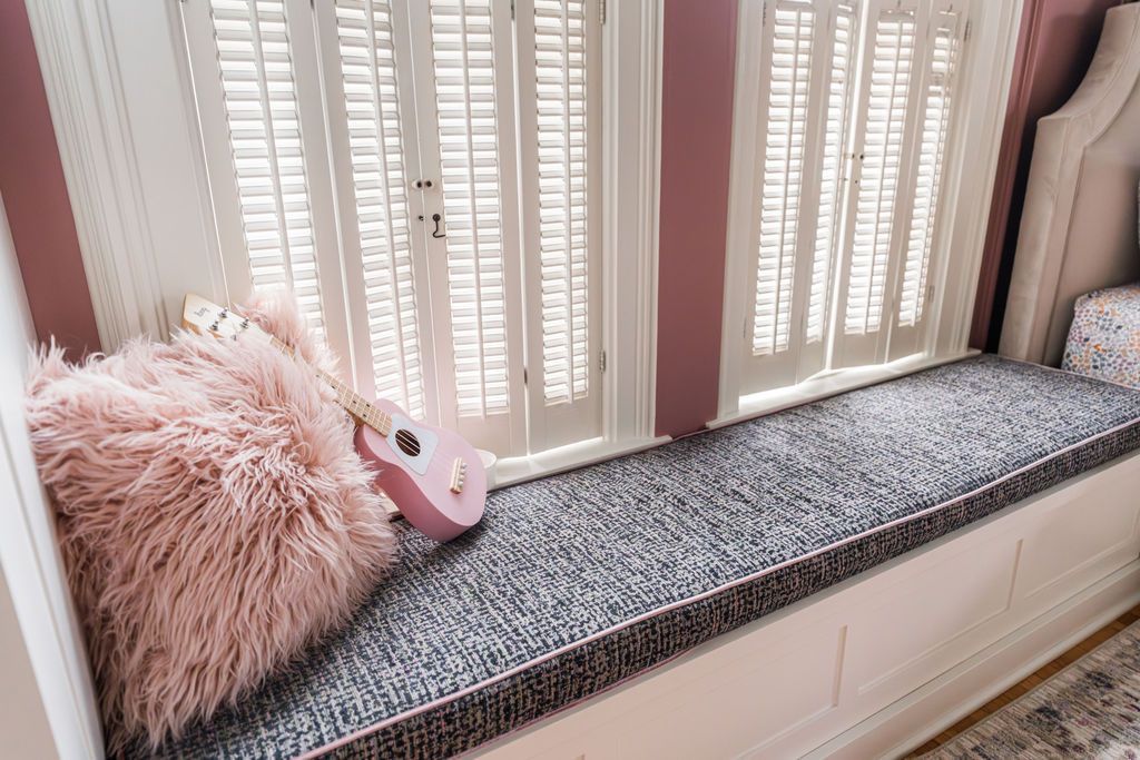 Cozy window seat with gray patterned cushion, pink fluffy pillow, and ukulele. White shutters and pink wall trim.