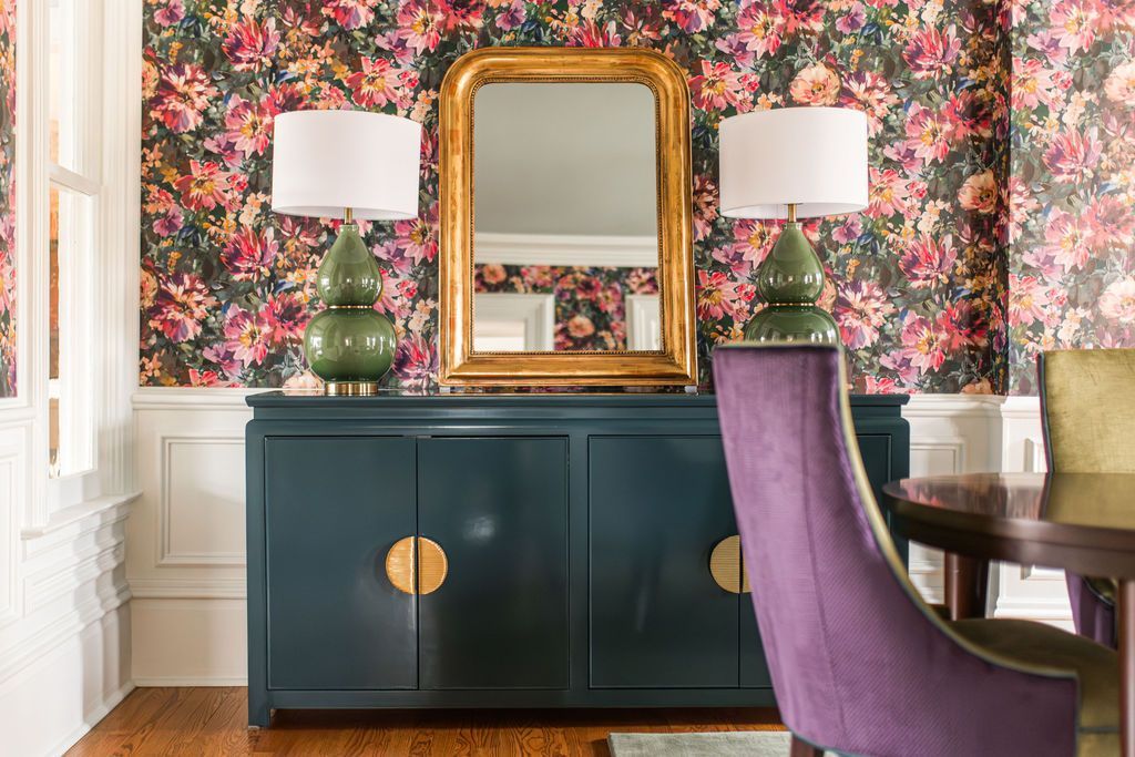 Dark blue cabinet with gold accents, mirror, lamps, and floral wallpaper in a dining room.