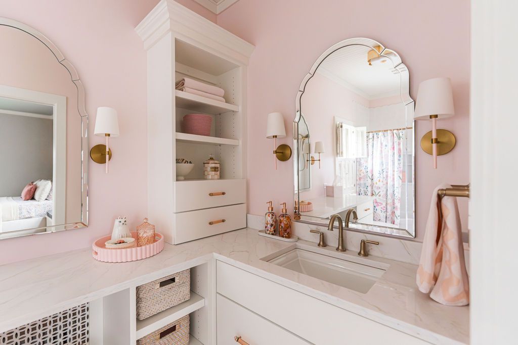 Pink bathroom with arched mirrors, white vanity, gold sconces, and built-in storage.