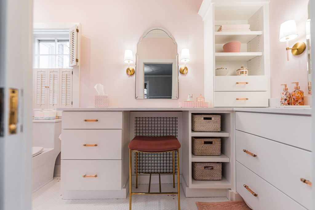 Pink and white vanity area with built-in storage, arched mirror, gold accents, and a patterned stool.