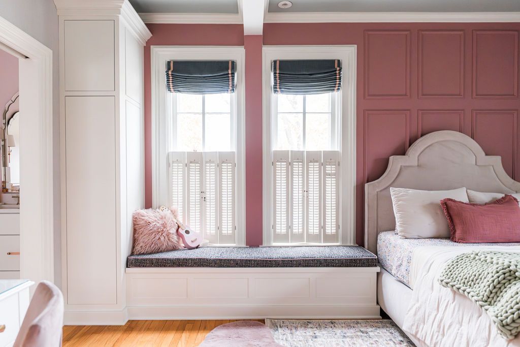 Bedroom with mauve walls, white shutters, a window seat, and a bed with a pink pillow and green throw.