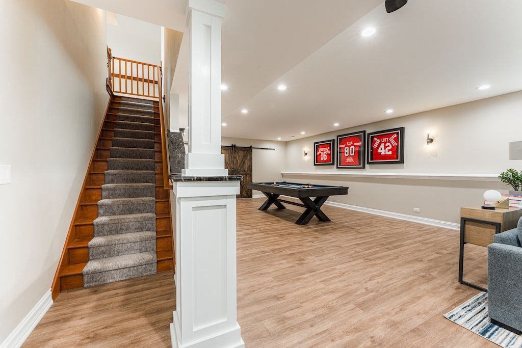 Basement recreation room with stairs, pool table, framed artwork, and wood-look flooring.