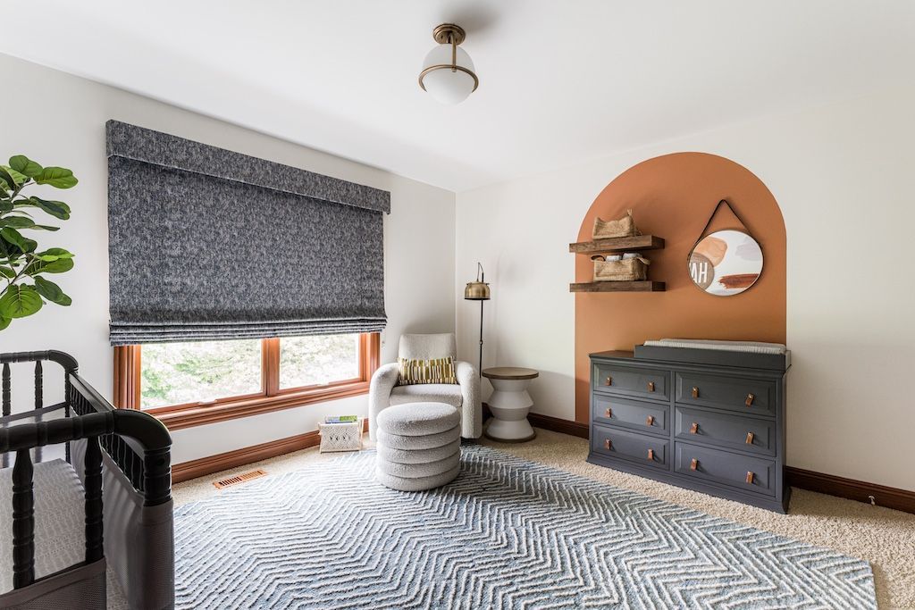 A nursery with a crib, changing table, and reading chair, featuring a modern rug and rust-colored arch.