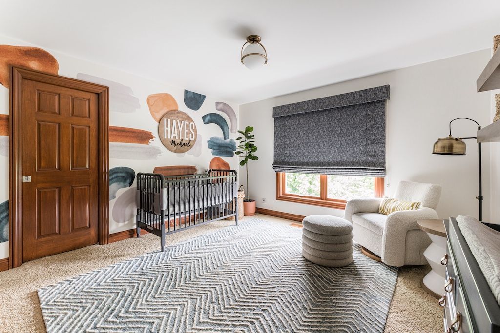 Nursery with a crib, armchair, and abstract wall art in a room with a patterned rug and wooden door.